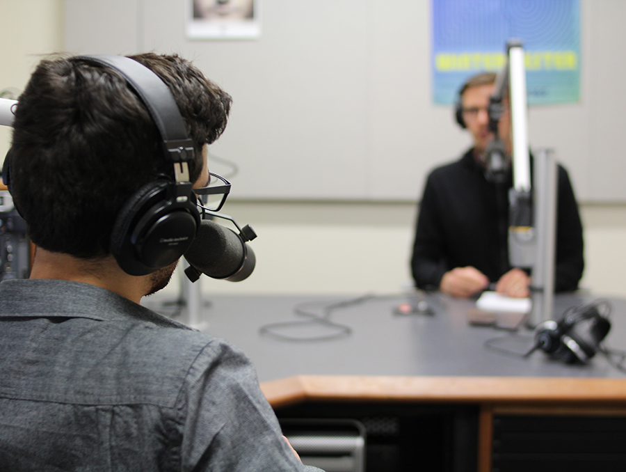 Two men sit across from each other in a podcasting studio. 