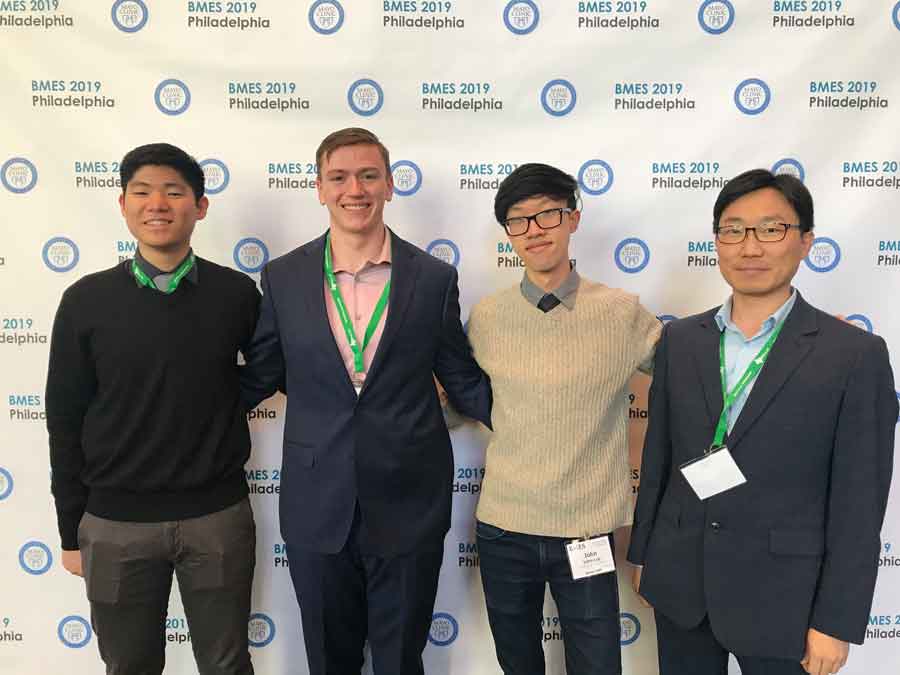 Three students and a professor stand in front of a conference wall.