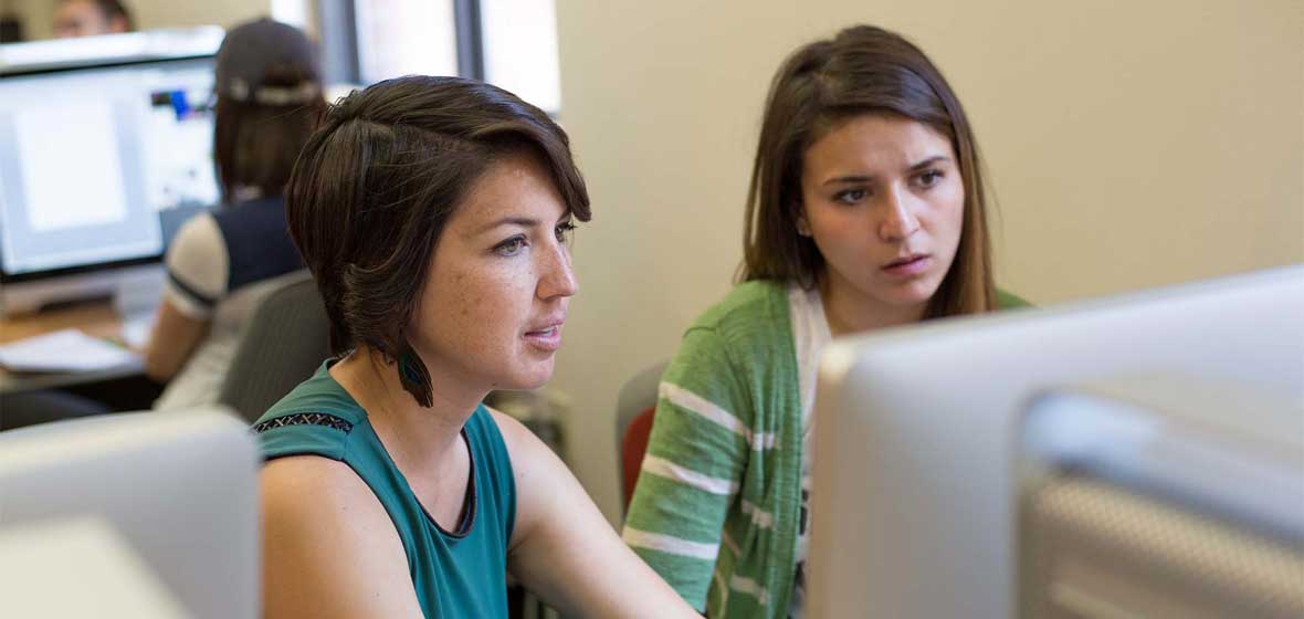 Two women sit in front of a computer. 