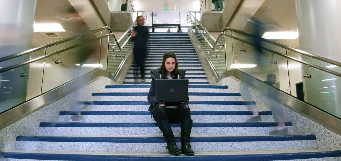 Unviersity student sitting on the stairs in the Ansari Business Building, working on laptop.