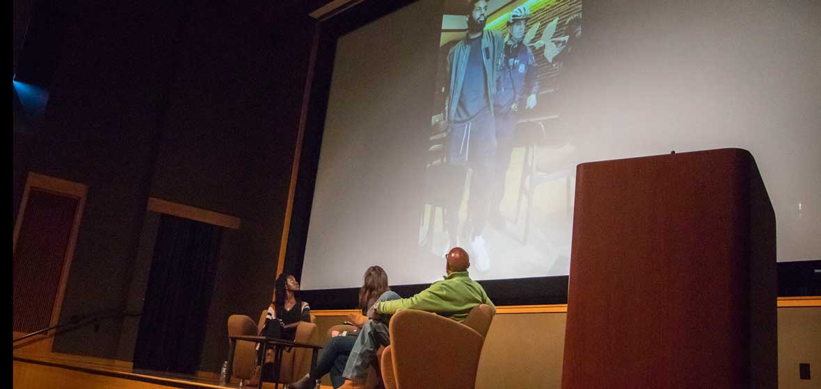 Three people on stage look up at a screen behind them. 