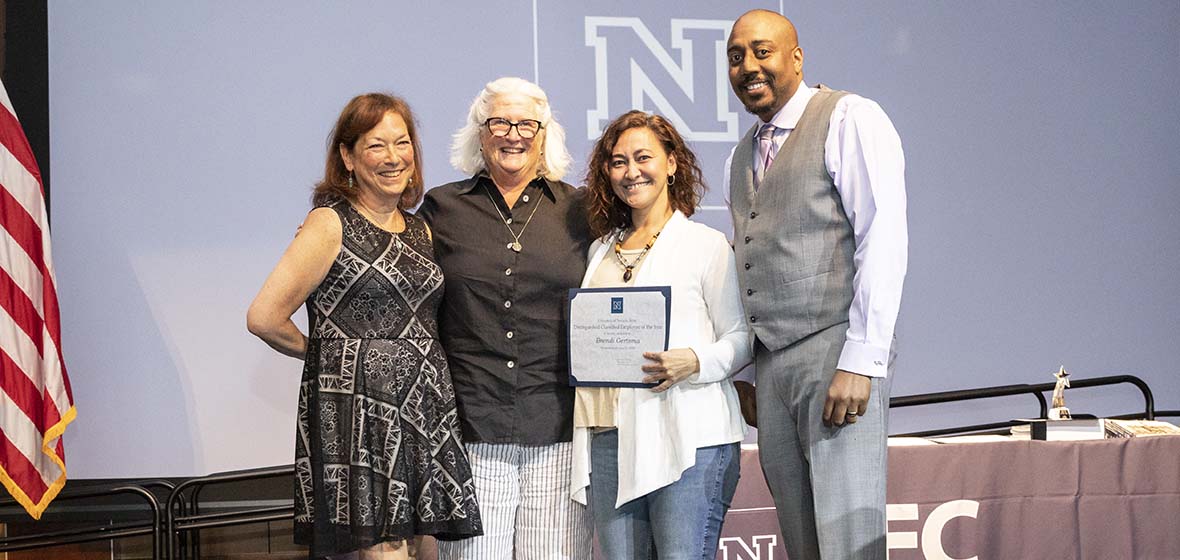 A portion of an American flag on the left with Donna Healy, Jacque Ewing-Taylor, Brendi Gertsma and Shawn Norman posing for a photo with one holding a certificate.