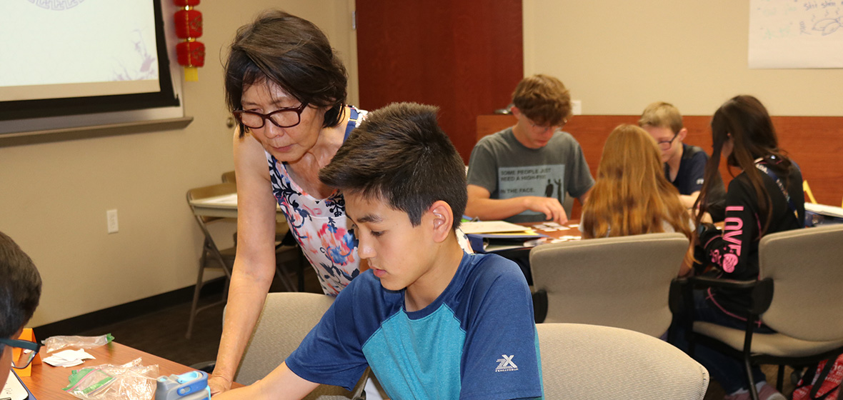 Chinese instructor leans over shoulder of young student to help with assignment at table