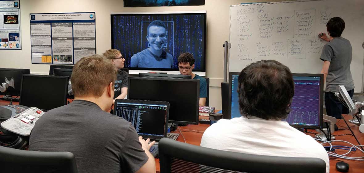 A group of students coding in a computer lab as one student writes instructions on a white board.