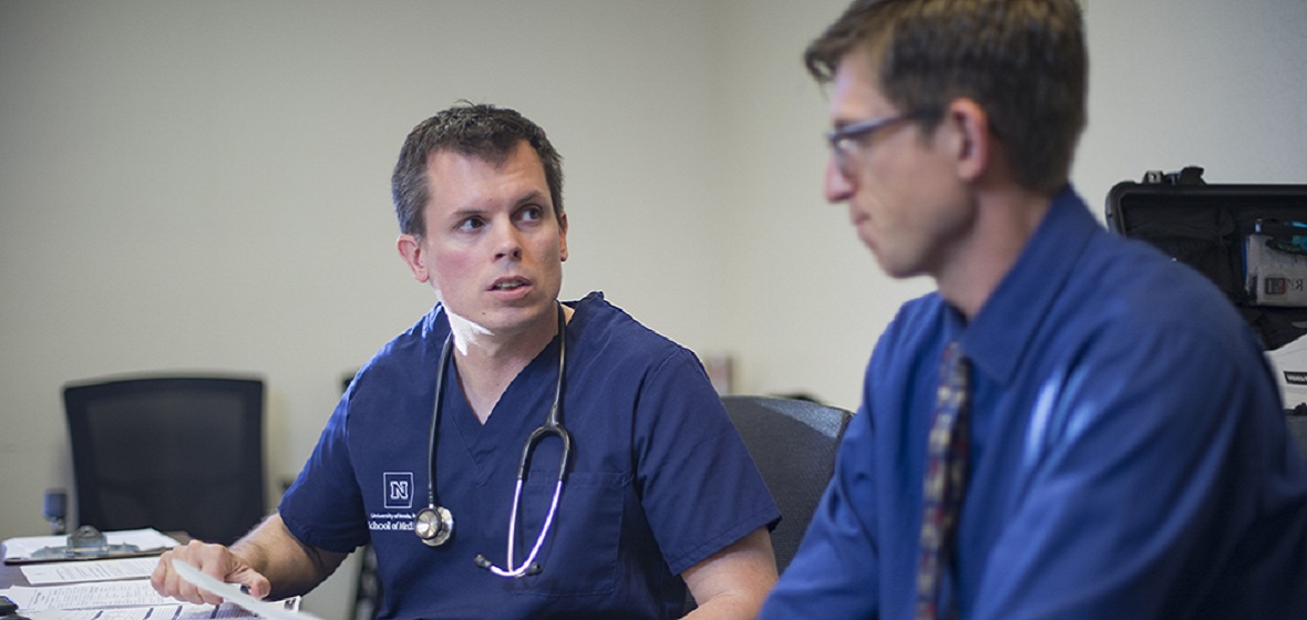 A male doctor in Nevada blue scrubs talks to a male patient
