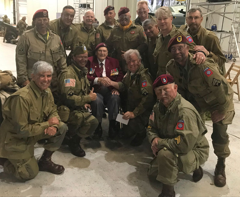 Group of US Army Veterans group around a WWII veteran for a photo.