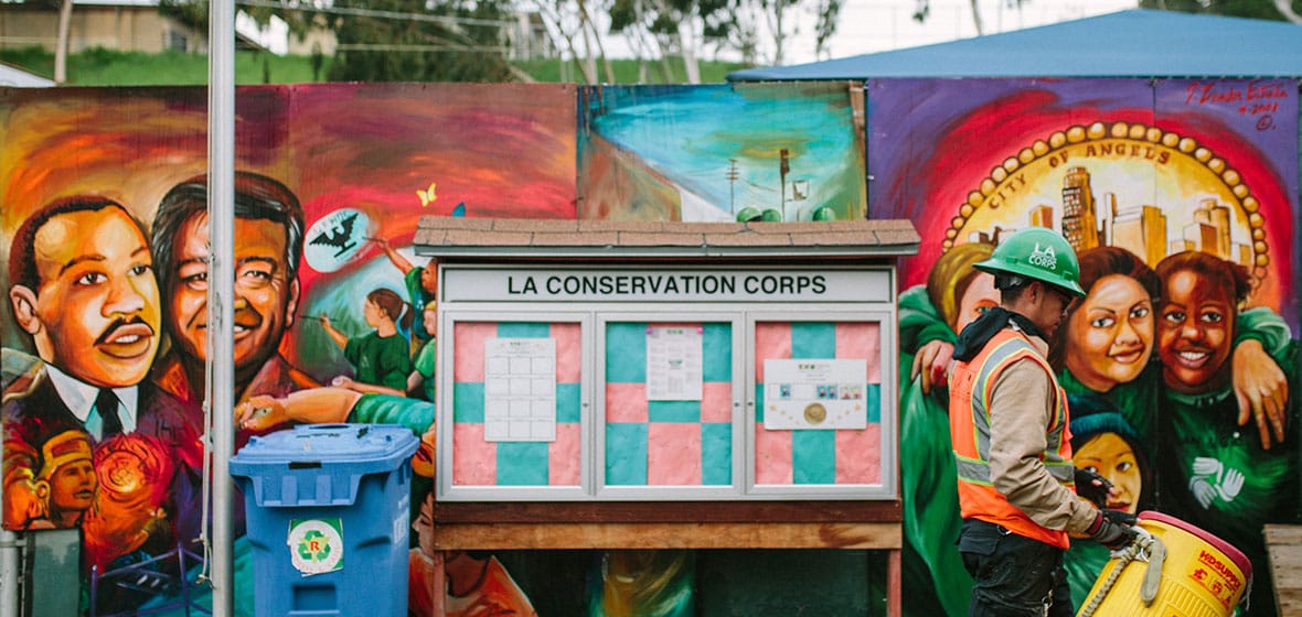 A man carries a water jug outdoors. Behind him is a mural and an announcement board with the title, LA Conservation Corps. 