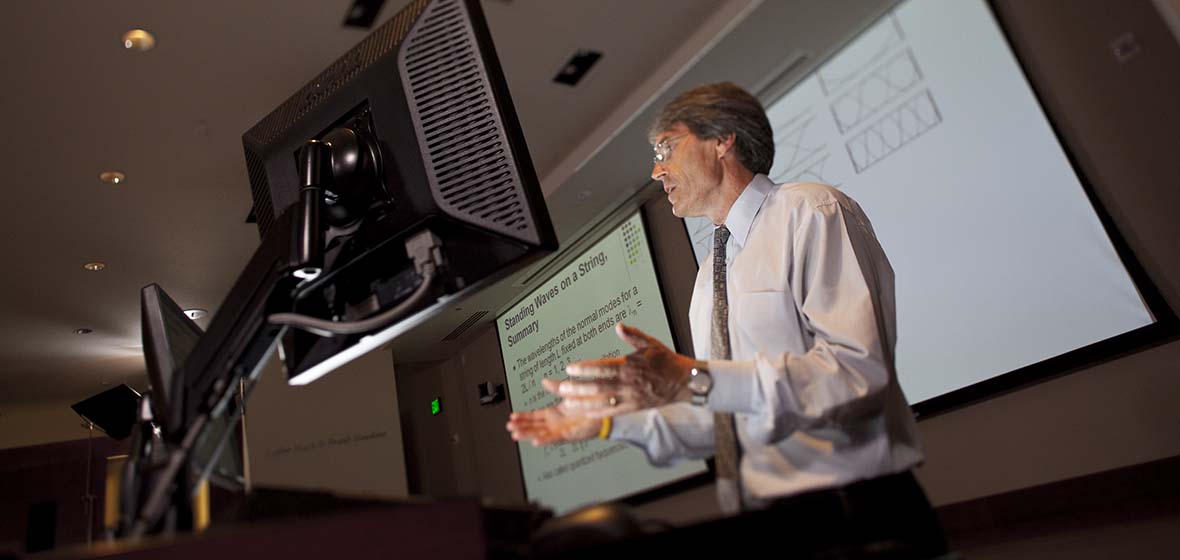 A professor stands behind a computer monitor during a lecture with a projector screen behind him.