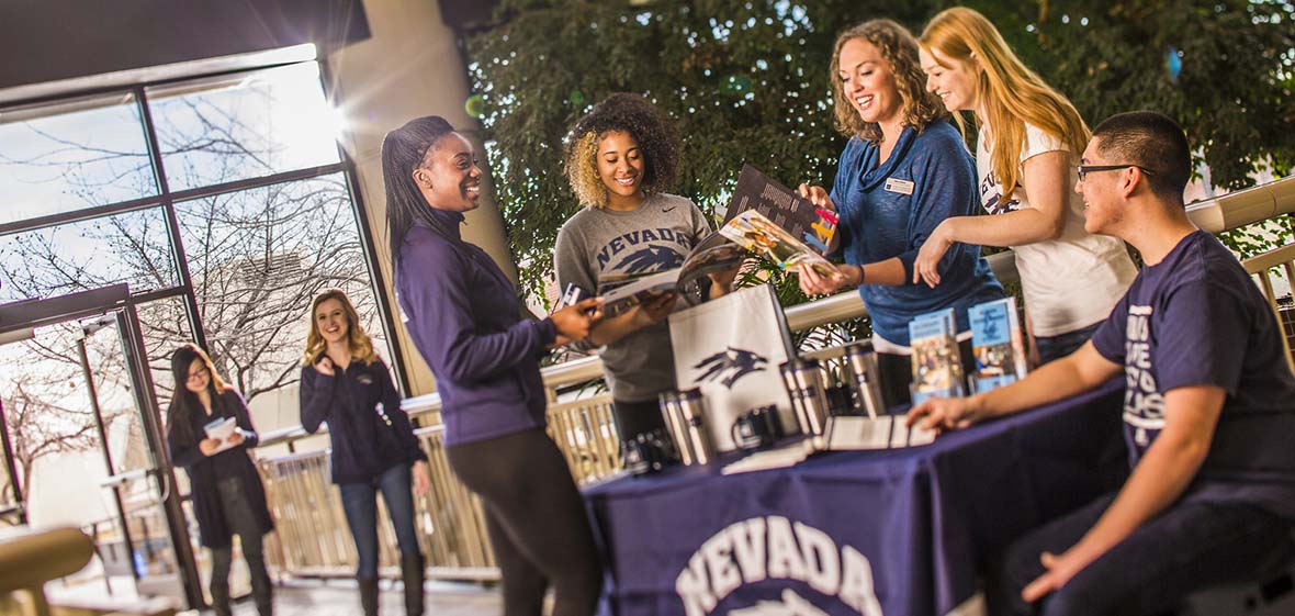 Two people enter a building and walk toward a table where five people are gathered around, one sitting, four standing, looking at brochures about the University of Nevada, Reno.