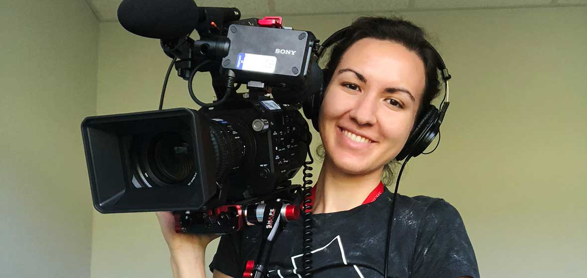 A woman smiles for the camera, holding a large camera over her shoulder.