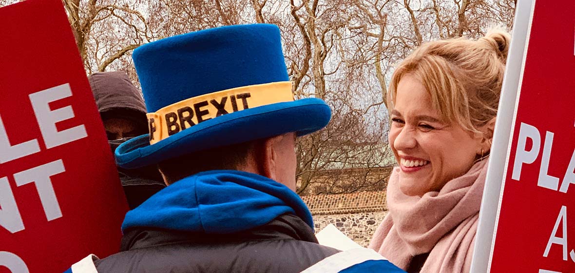 A person with a blue and yellow BREXIT top hat looks at another person smiling, wearing a light pink scarf. Red and white signs with nondescript words are on either side of the two people. 