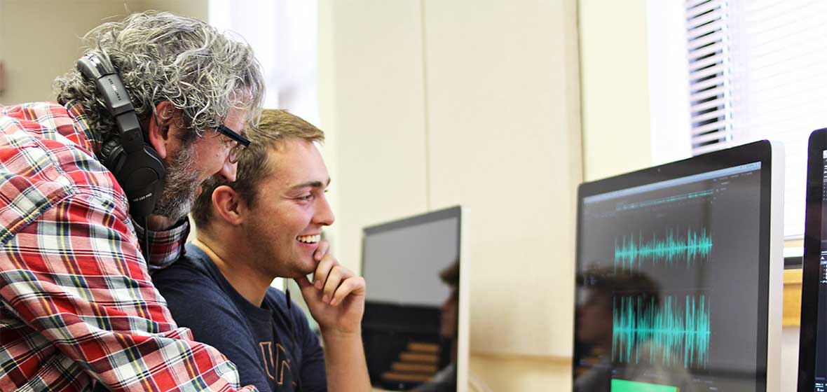 A professor leans over a student's shoulder while they both look at a computer screen. 