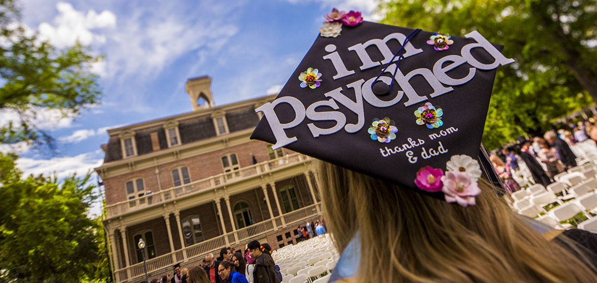 Graduate facing Morril Hall with a mortar board that reads, "I'm psyched. Thanks Mom & Dad"