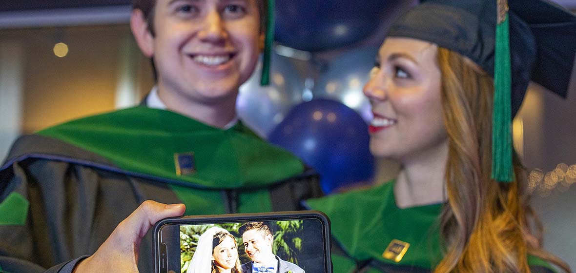 Two graduates pose in their commencement regalia, holding a cell phone displaying a photo with the two of them dressed as bride and groom on their wedding day