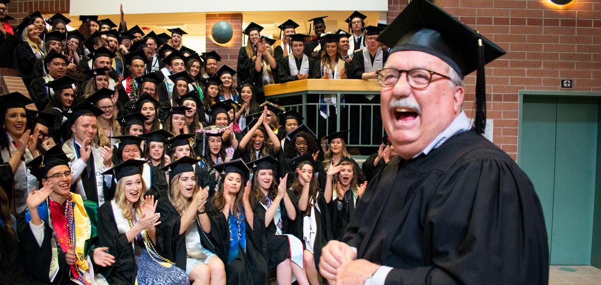 A man in graduation robes smiles before a large group of students. 