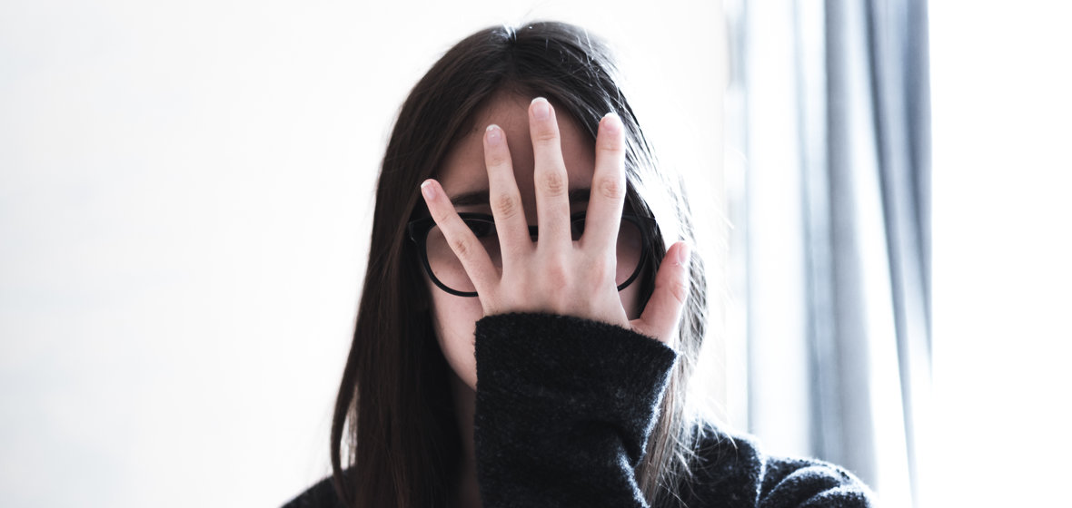 Woman with dark hair, a black sweater, and glasses hiding her face behind her hand