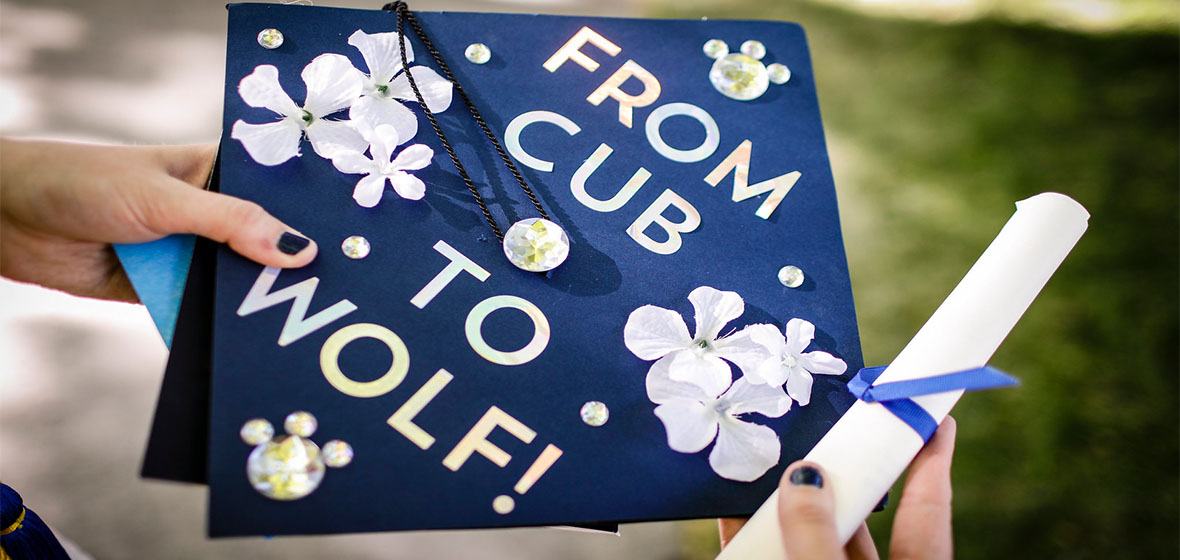 A person holding a degree and a graduation cap that says From Cub to Wolf 