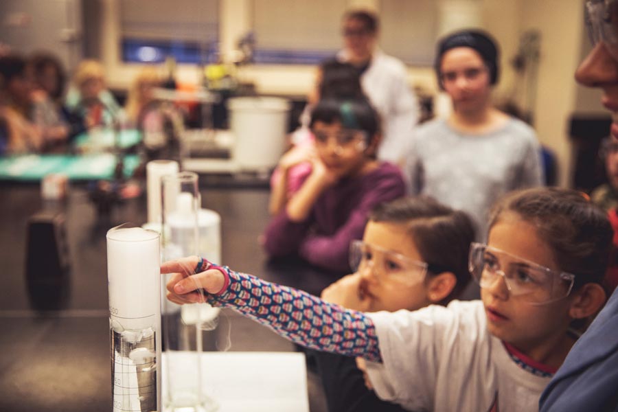A young girl wearing safety goggles reaches out to touch the gas spilling out of a beaker.