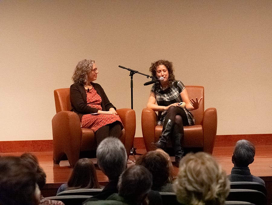 A woman speaks into a microphone on stage with another woman sitting nearby.