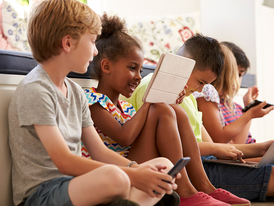 Five children sitting in a row playing on various tech.