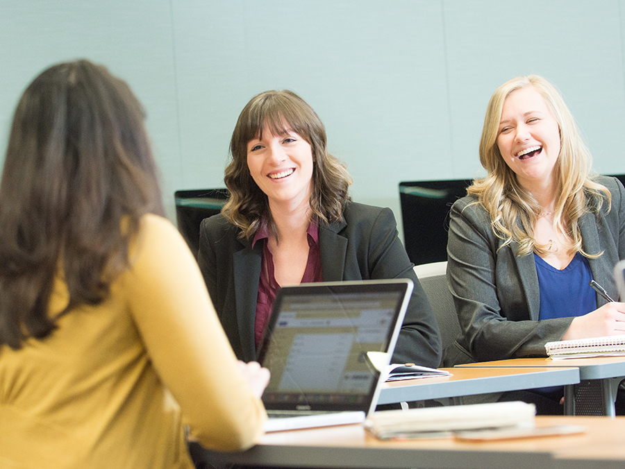 Three women sit in a computer lab and laugh together.