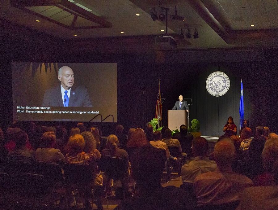 President Marc Johnson speaks at the podium during the State of the University Address. A live video of him speaking is playing to his right, and the caption reads, 'Higher Education rankings. Wow! The University keeps getting better in serving our students!'