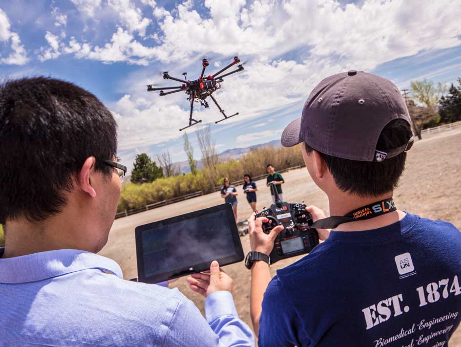 A drone hovers in the air while two researchers, seen from the back, look on with an iPad and camera