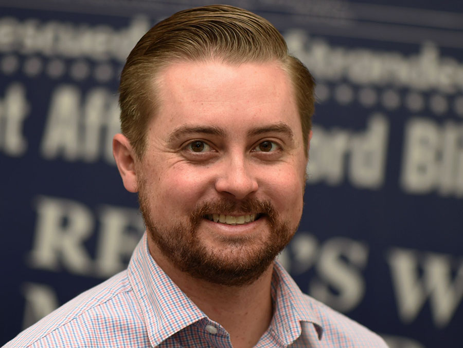 Brian Duggan is pictured smiling in front of a blue background smiling while wearing a light blue and red plaid button up.