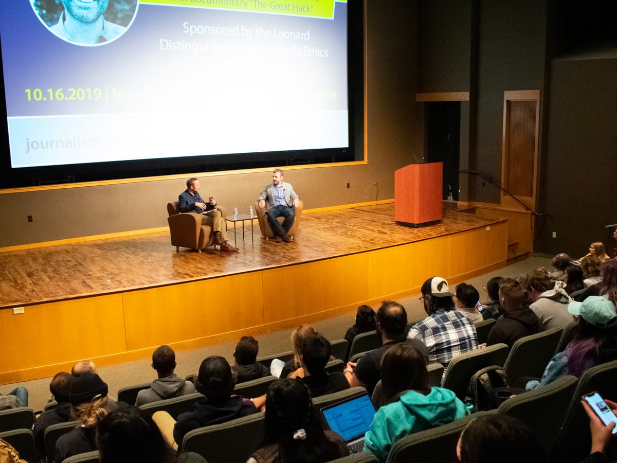 Two men sit on stage as an audience watches.