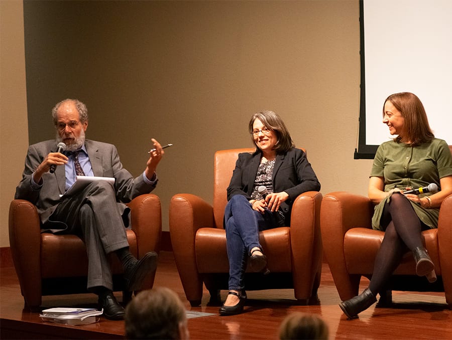 A man speaks while two woman listen on stage.