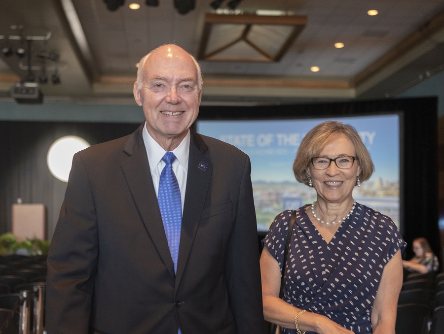 President Marc Johnson with wife Karen Penner-Johnson, a slide with a shot of the University and the words State of the University can be seen in the background