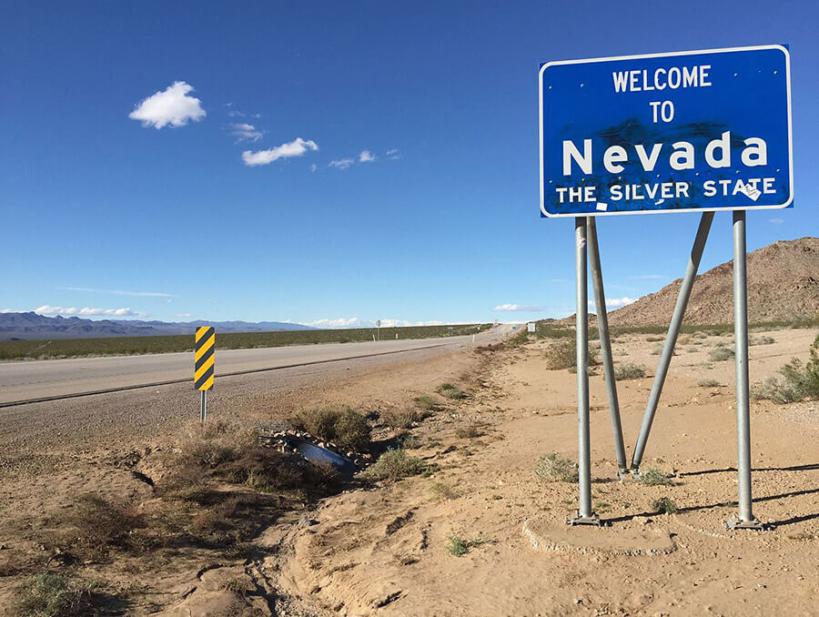 Landscape of a desert highway with a blue road sign that says "Welcome to Nevada, The Silver State" 