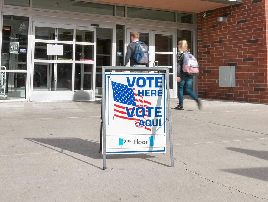 Sign saying "vote here Vote aqui" outside the Joe Crowley Student Union, with two students walking in