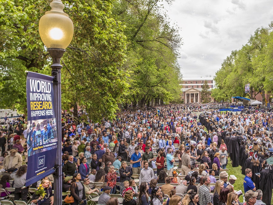 Spring Commencement on the Quad. A line of graduates in robes make their way through a crowd of family and friends. A light pole with a banner reading 'World Improving Research Happens Here' is in the foreground.