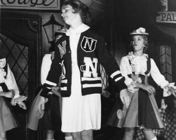 The Homecoming Wolves Frolic skits sorority competition shows a scene with a girl in a cheerleader jacket and white dress in the foreground with three girls in costumes in the background. (1961)