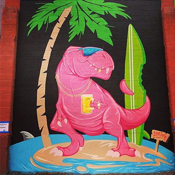 Elko Mural Expo mural by Reno-based muralist Mike Lucido. This mural features a Pink T-Rex dinosaur holding a green surfboard