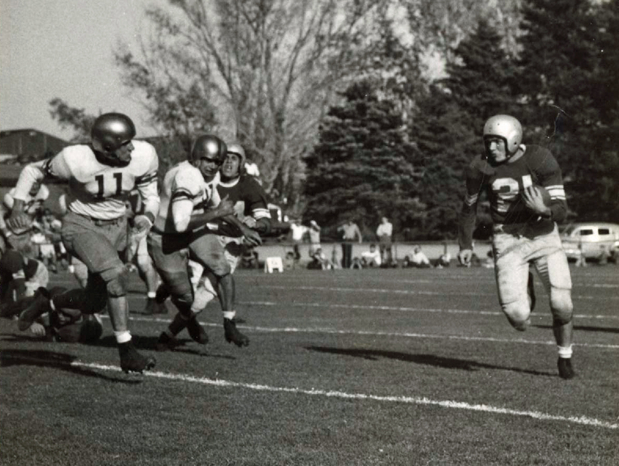 Black and white image of men in football uniform chasing another player holding the ball.