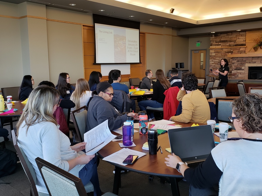 A conference room with about 25 students and faculty sitting around several tables and watching a facilitator lead a presentation on a large screen.