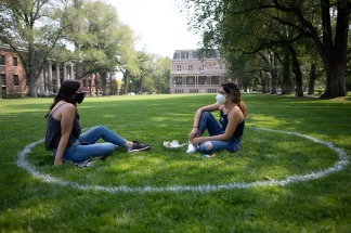 Two students sitting with masks on in a spray-painted white circle on a grass lawn