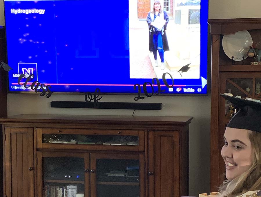 Student sits in living room with a cap and gown on with the University of Nevada, Reno commencement program on the television.