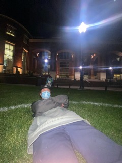 A student lays on the lawn with as mask on in front of the knowledge center in a white circle
