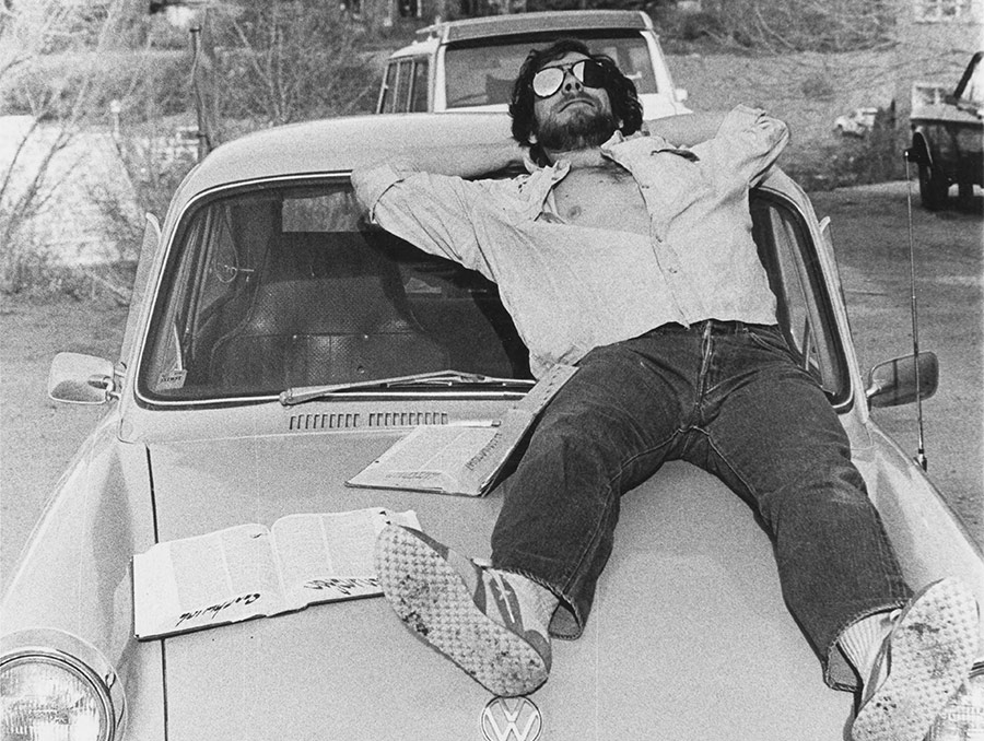 Black and white  photograph of a man wearing sunglasses and laying on top of a white car, 1979