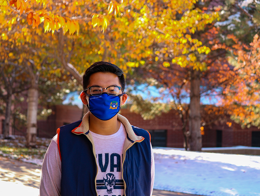 Student standing in front of trees with fall leaves and they have a mask on