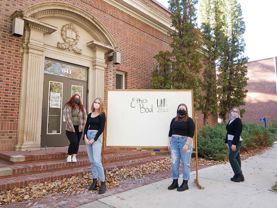 Four Ethics Bowl team members pose outside with a whiteboard showcasing their team name