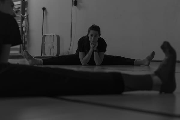 A black and white image of Keely Cobb siting in a wide straddle stretch with her legs spread out, her elbows on the ground and her head resting on her hands in a dance studio.
