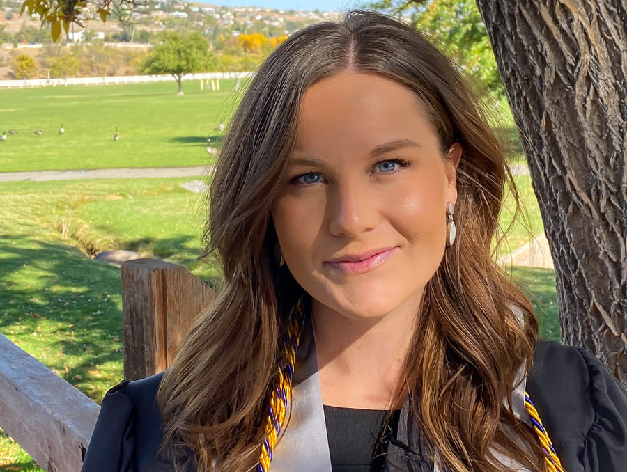 A headshot of Tamara Snight standing in front of a park wearing a black graduation gown and smiling to camera.