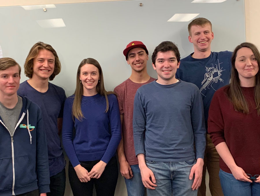 A group of students and their professor pose in front of a dry erase board.