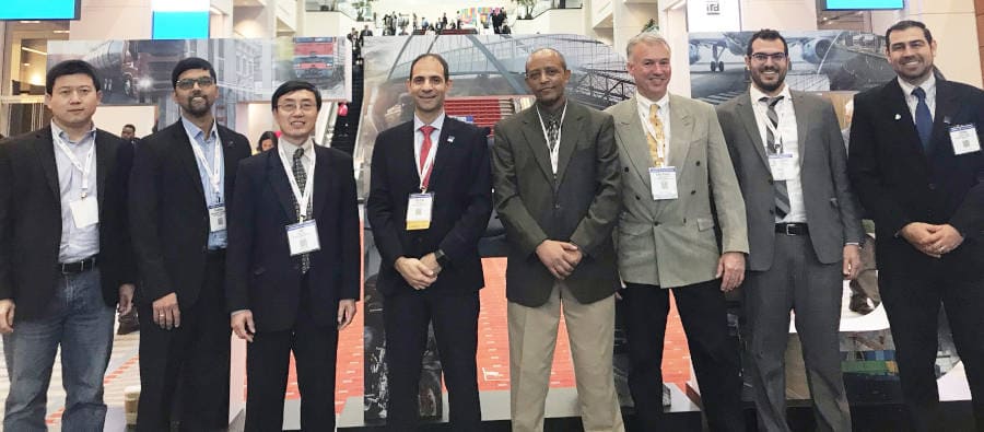 Eight men in business attire, members of the pavement project team, pose for a group photo in an exhibition hall. 