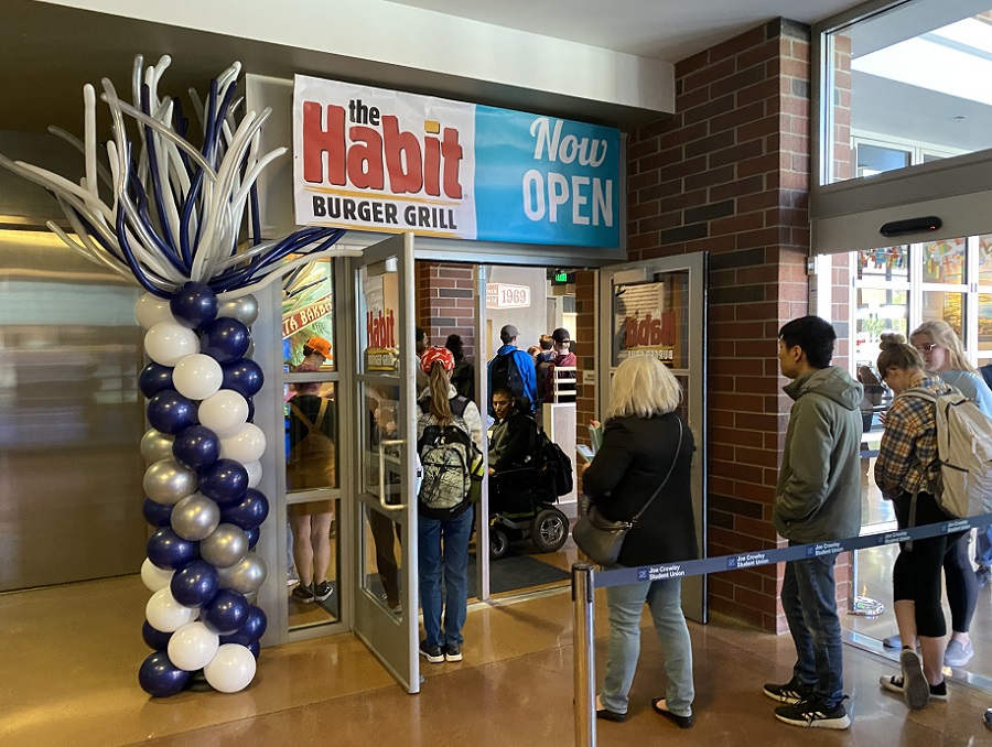 The line at the opening of Habit Burger Grill. Students are inside the location and entering under a "the Habit Burger Grill - Now open!" sign and a silver and blue balloon palm tree.