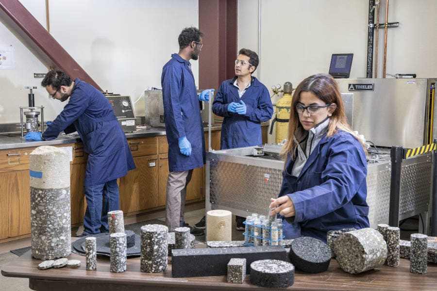 Grad students, three men and one woman, in blue lab coats work in a lab surrounded by pavement samples and related materials.